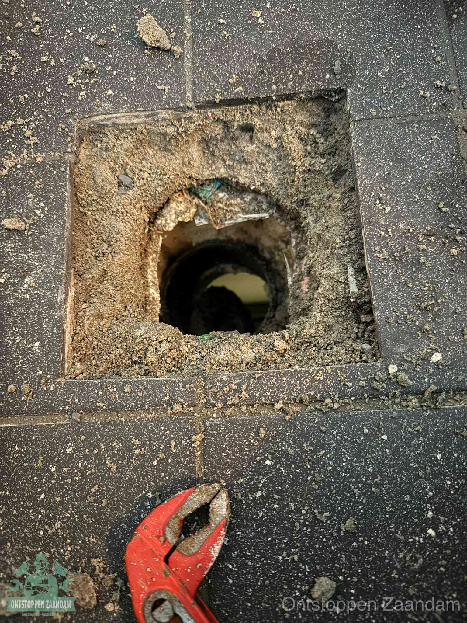 Een geopend rioolputje in een betontegelvloer met losgemaakt rooster en een zichtbare ronde afvoerbuis, het achterhalen waar de afvoerverstopping zit in Zaandam