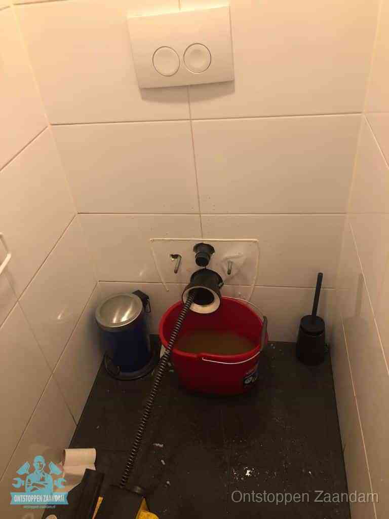 Een ontstopte wand-wc met verwijderde toiletpot waar een ontstoppingsveer in de wanduitgang verdwijnt, het achterhalen waar de verstopping zit in Zaandam