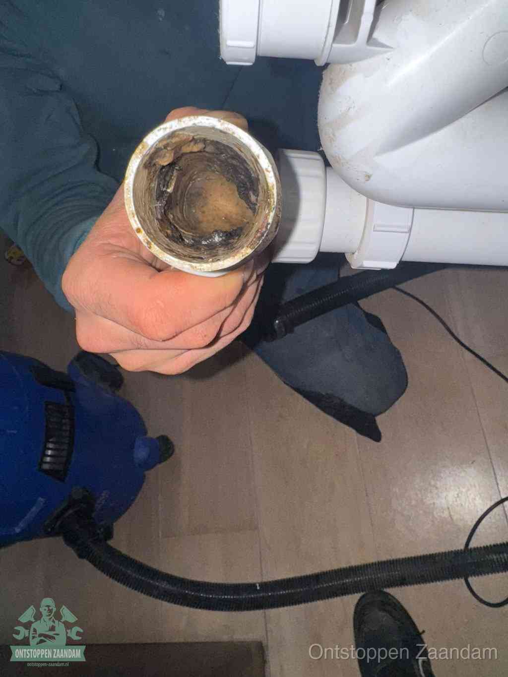Een hand houdt een losgekoppeld PVC-afvoerbuisstuk vast dat van binnen volledig is dichtgeslibd met vet, vuil en aanslag, de meest voorkomende oorzaak van een verstopte afvoer in Zaandam