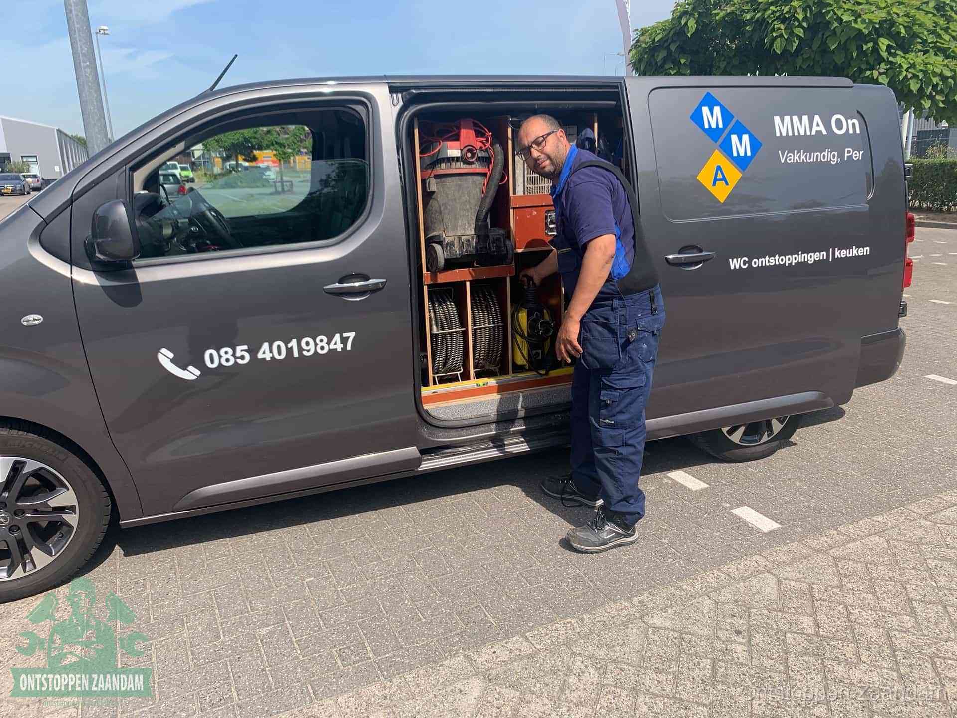 Een monteur bij de open schuifdeur van zijn bedrijfsbus op een parkeerplaats, klaar om de oorzaak van terugkerende afvoerverstoppingen aan te pakken in Zaandam