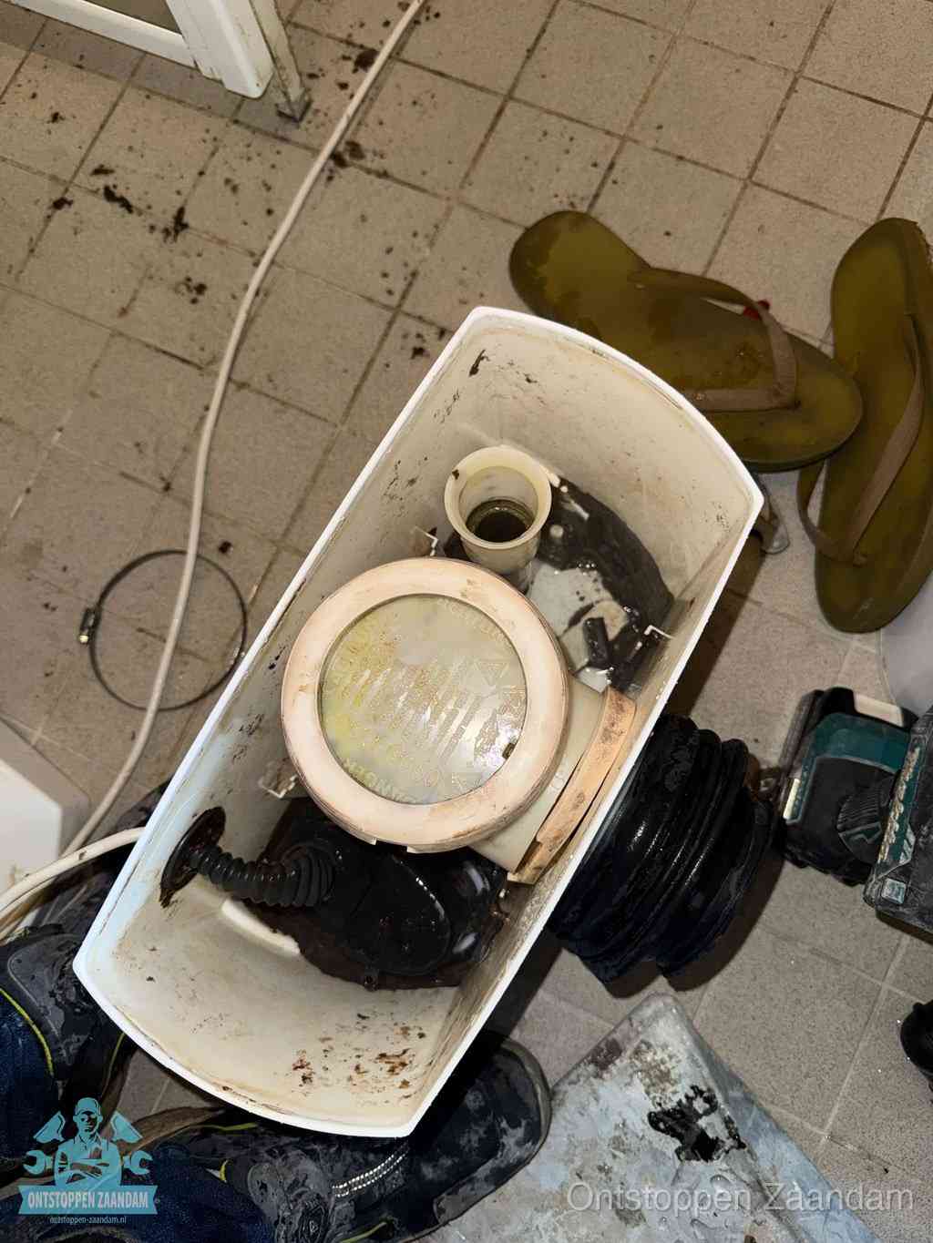 Een gedemonteerde toiletpot op de vloer met ontstoppingsveer en gereedschap ernaast, het verwijderen van een vastzittend voorwerp uit de wc in Zaandam