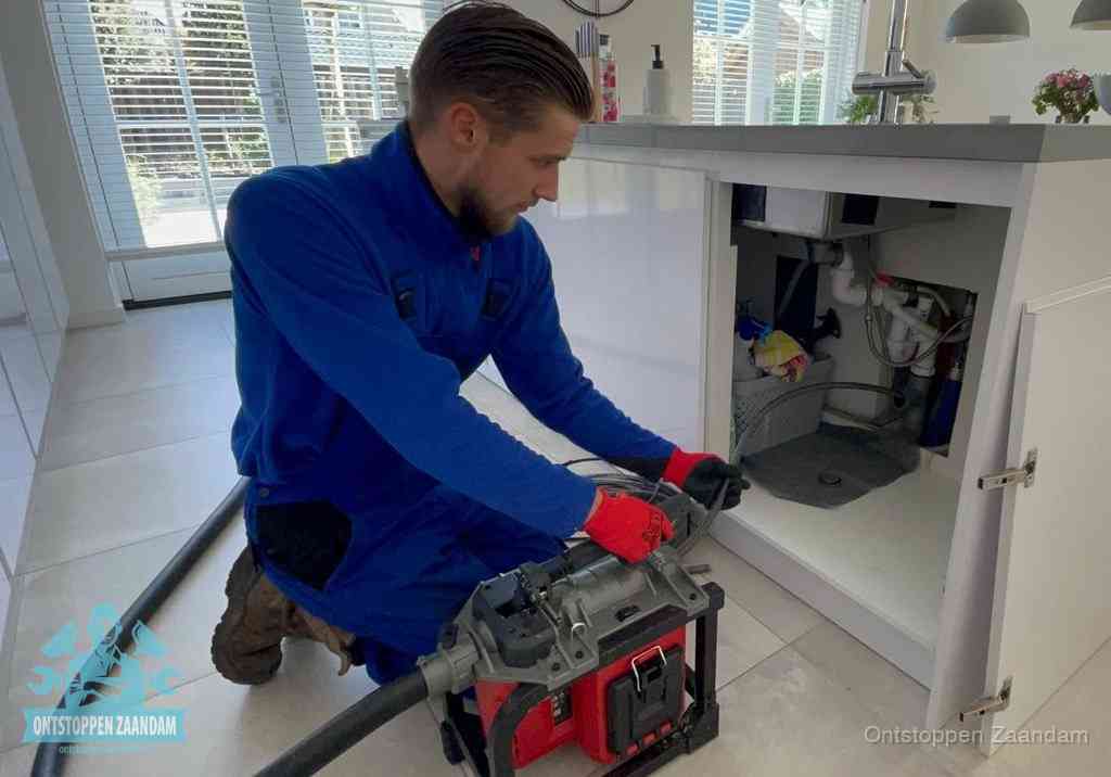 Een monteur voert een professionele elektrische ontstoppingsmachine in de afvoer onder de spoelbak tijdens het gootsteen ontstoppen in Zaandam